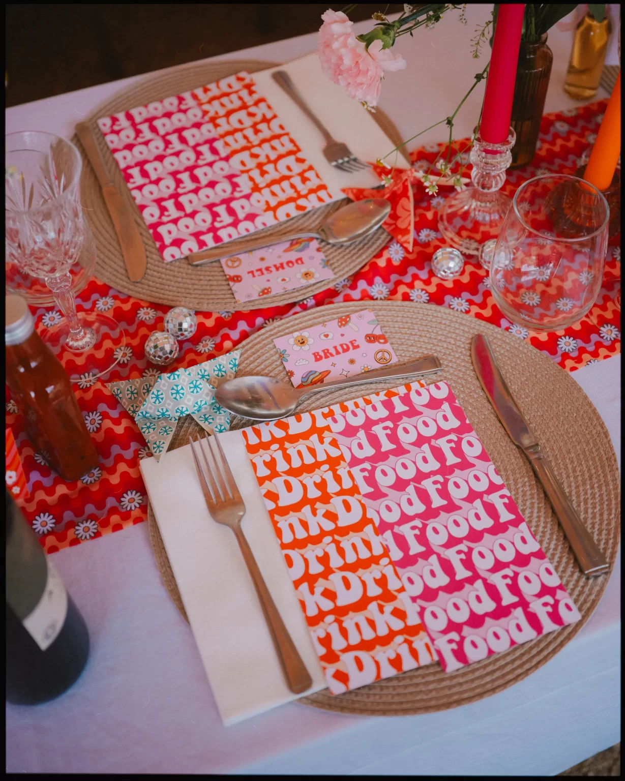 A colorful table setting with plates, napkins reading “Drink Food” in bold letters, cutlery, glasses, and vibrant candles on a red patterned table runner. A small card labeled “BRIDE” rests near one plate.