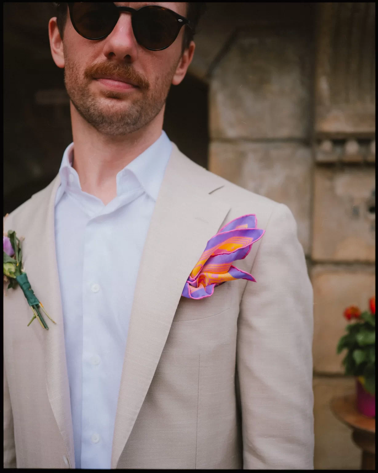 A man in a light beige suit with a white shirt wears sunglasses and has a colorful purple and orange pocket square. Flowers are pinned to his lapel, and blurred flowers and stone architecture appear in the background.