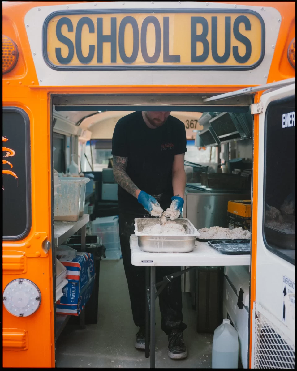 A person wearing gloves prepares food at a table set up inside a converted school bus, with kitchen equipment visible in the background and the word SCHOOL BUS displayed above.