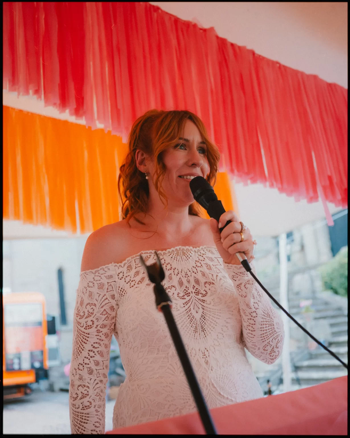 A woman in an off-the-shoulder white lace dress speaks into a microphone under red and orange streamers, smiling with wavy hair tied back, at what appears to be a festive event at guys cliffe house, warwick