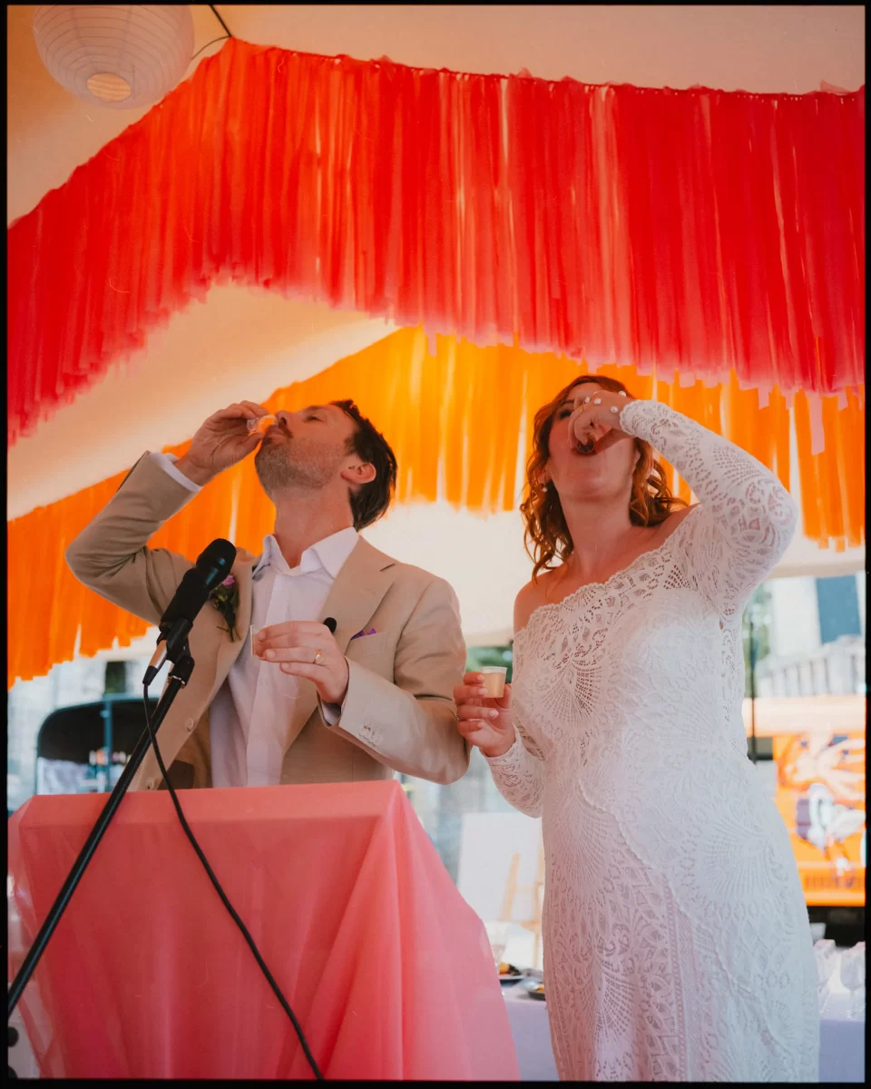 A couple in wedding attire stands under an orange and pink canopy at guys cliffe house, warwick, taking a drink together at a podium decorated with pink fabric and a microphone.