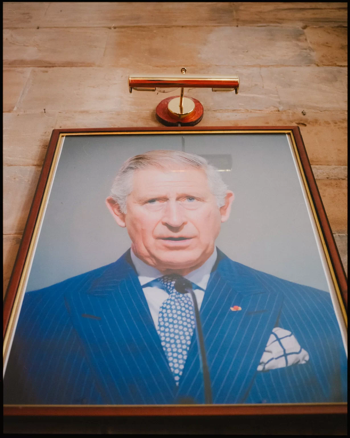 A framed portrait of an older man in a blue suit, patterned tie, and pocket square is mounted on a stone wall beneath a picture light.