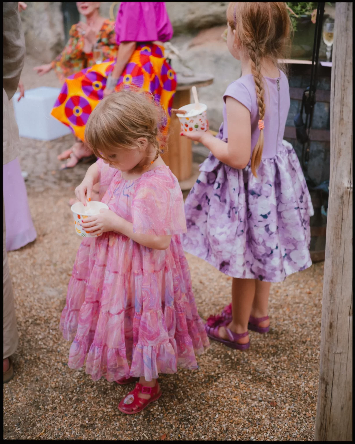 Two young girls in colorful, patterned dresses and sandals stand on a gravel surface at guys cliffe house, warwick, each holding a cup of ice cream. Adults sit in the background, dressed in bright clothing.