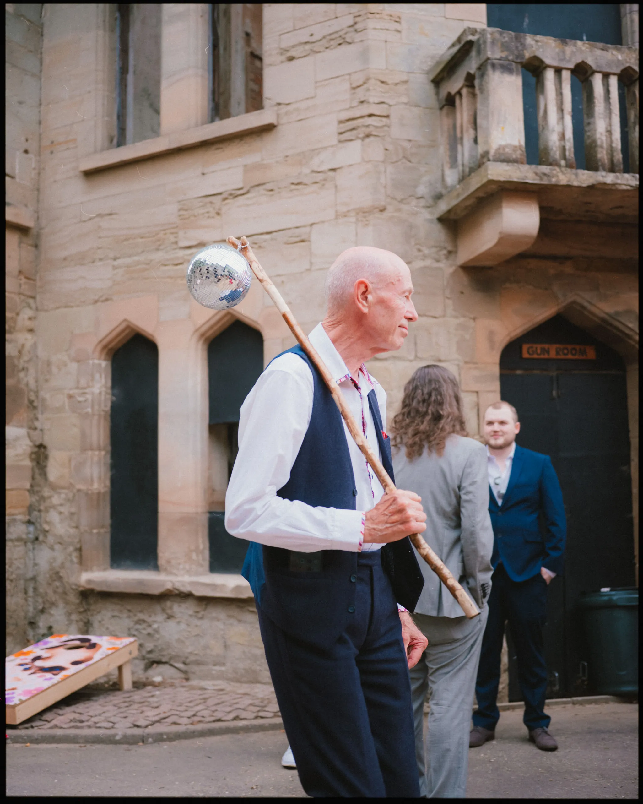 An older man in a vest and white shirt holds a staff with a disco ball on top. He stands outside a stone building, with two other people and a “Gun Room” sign visible in the background.