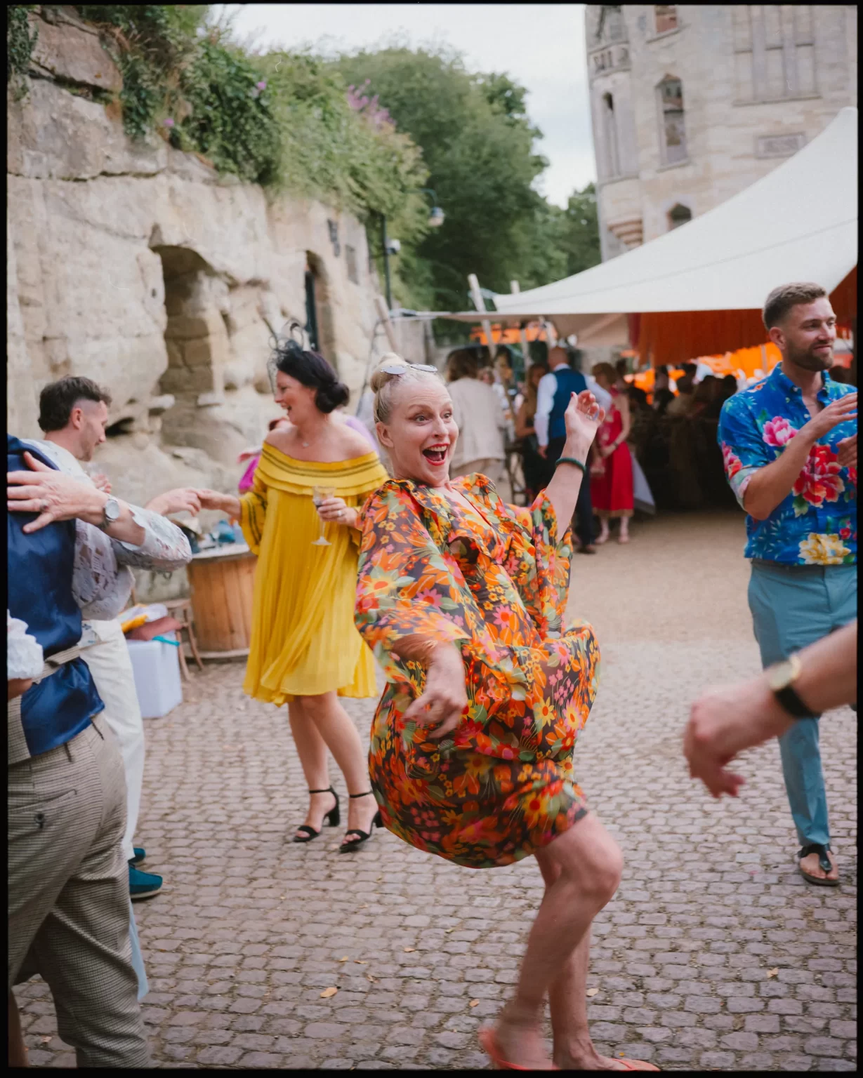 A joyful woman in a colorful floral dress dances enthusiastically at an outdoor event at guys cliffe house, warwick, surrounded by other people in bright outfits, with stone walls and a white tent in the background.