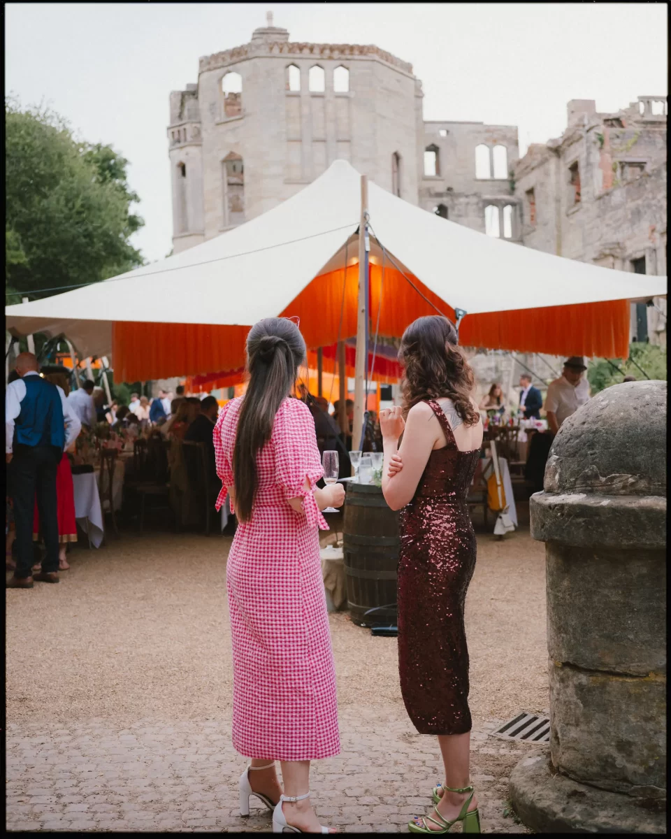 Two women in dresses holding drinks stand outside a large white tent at an event at guys cliffe house, warwick, with historic stone ruins and trees in the background. The tent glows warmly inside and guests mingle nearby.