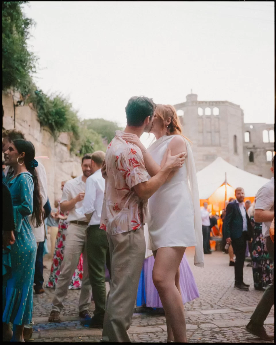 A couple shares a kiss while dancing outdoors at a festive event at guys cliffe house, warwick, surrounded by other guests in colorful outfits. Historic stone ruins and a white tent are visible in the background.