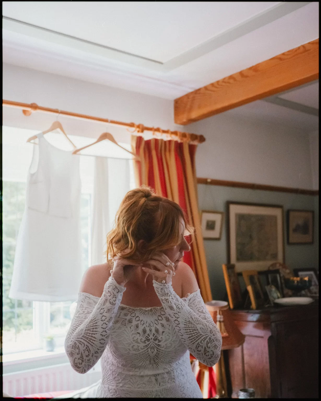 A woman in a white lace dress adjusts her earring in a warmly lit room, with a sleeveless dress hanging by the window and framed pictures on a wooden dresser behind her.