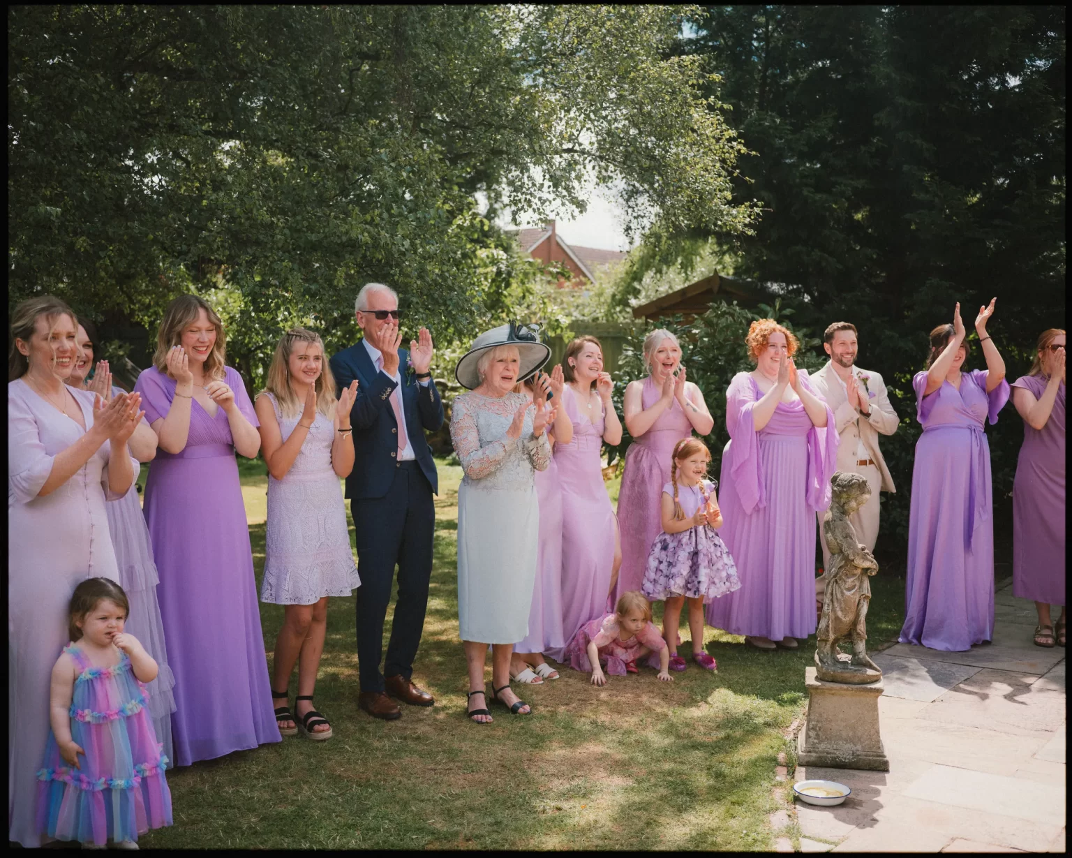 A group of people, mostly women and girls in matching pastel purple dresses, stand outdoors on grass, smiling and clapping. A few young children are in front, and trees provide shade in the background.