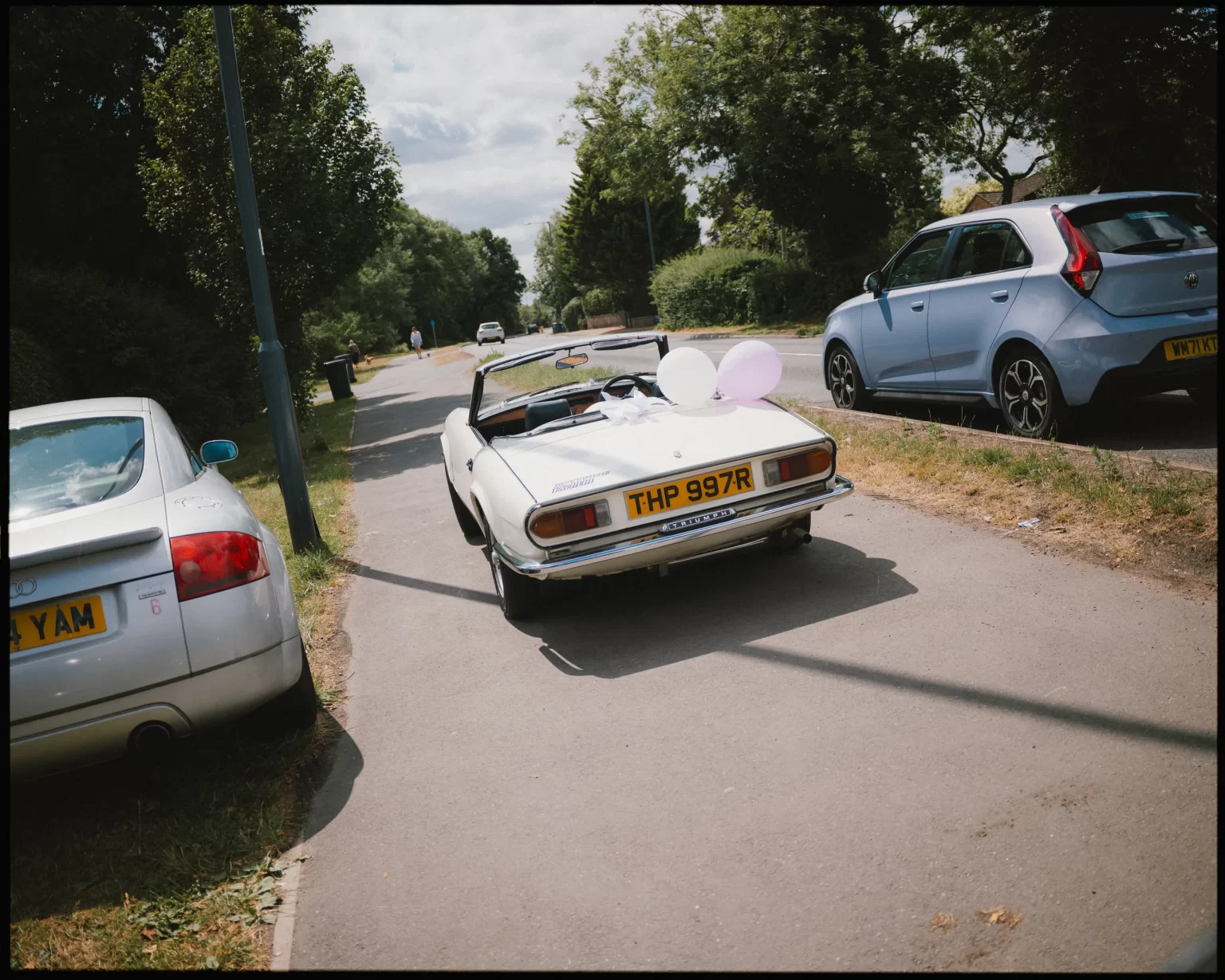 A white vintage convertible decorated with pink balloons is parked on the side of a sunny suburban street, surrounded by trees and other parked cars.