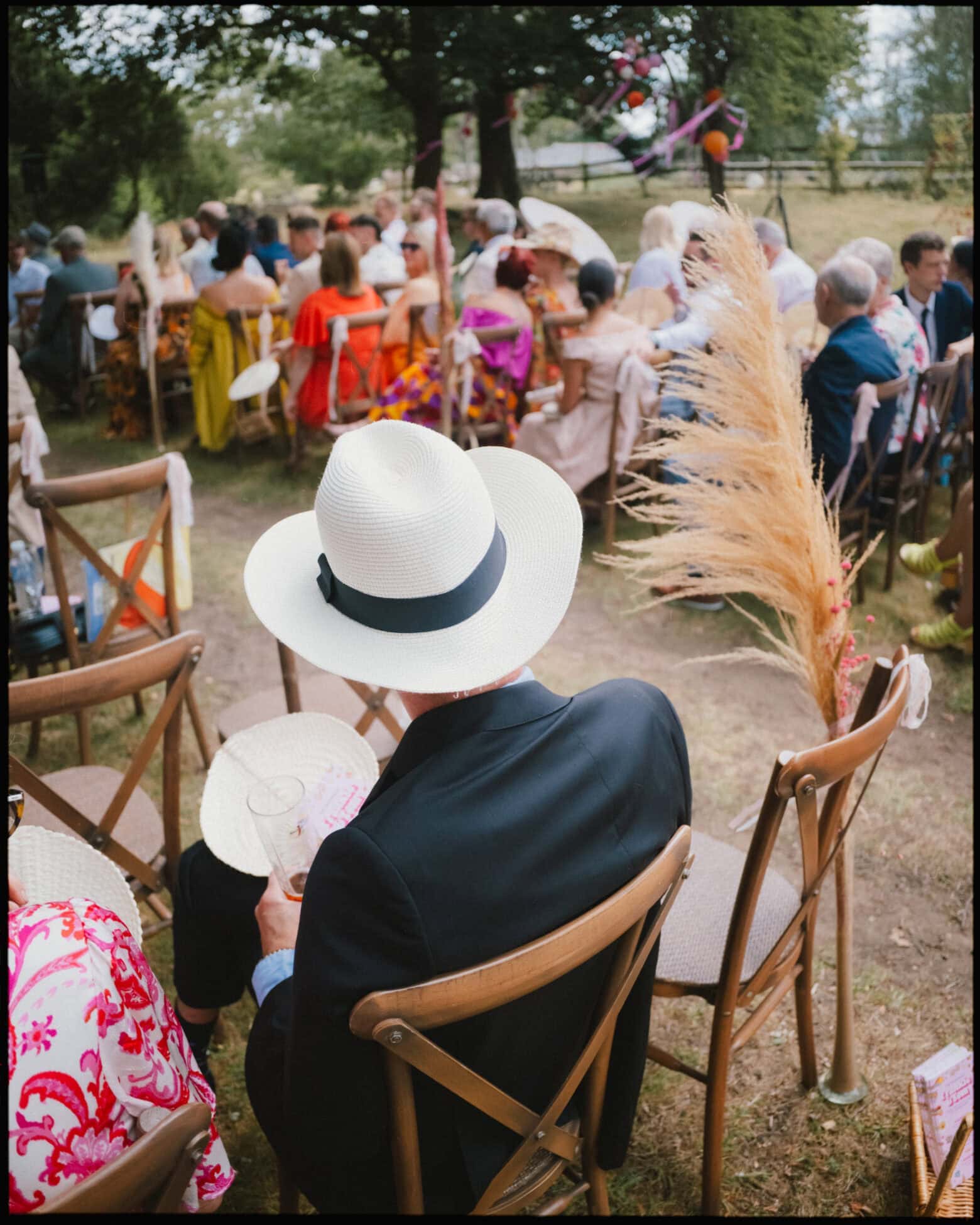 Wedding guest in a sun hat sitting at the outdoor ceremony with dried flower decorations at Guy’s Cliffe House in Warwickshire.