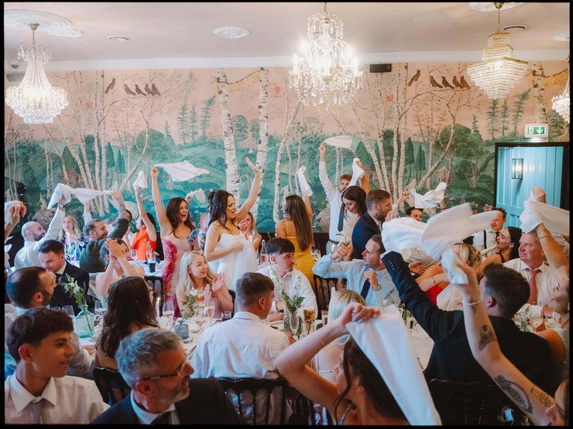 Guests cheering and waving napkins as the bride and groom make their entrance into the wedding breakfast at The Bell in Ticehurst.