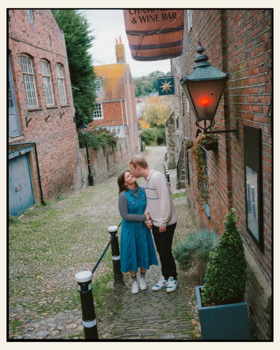 american couple walking up a cobbled narrow street with lantern in the ancient town of rye for their engagement shoot ahead of their destination wedding