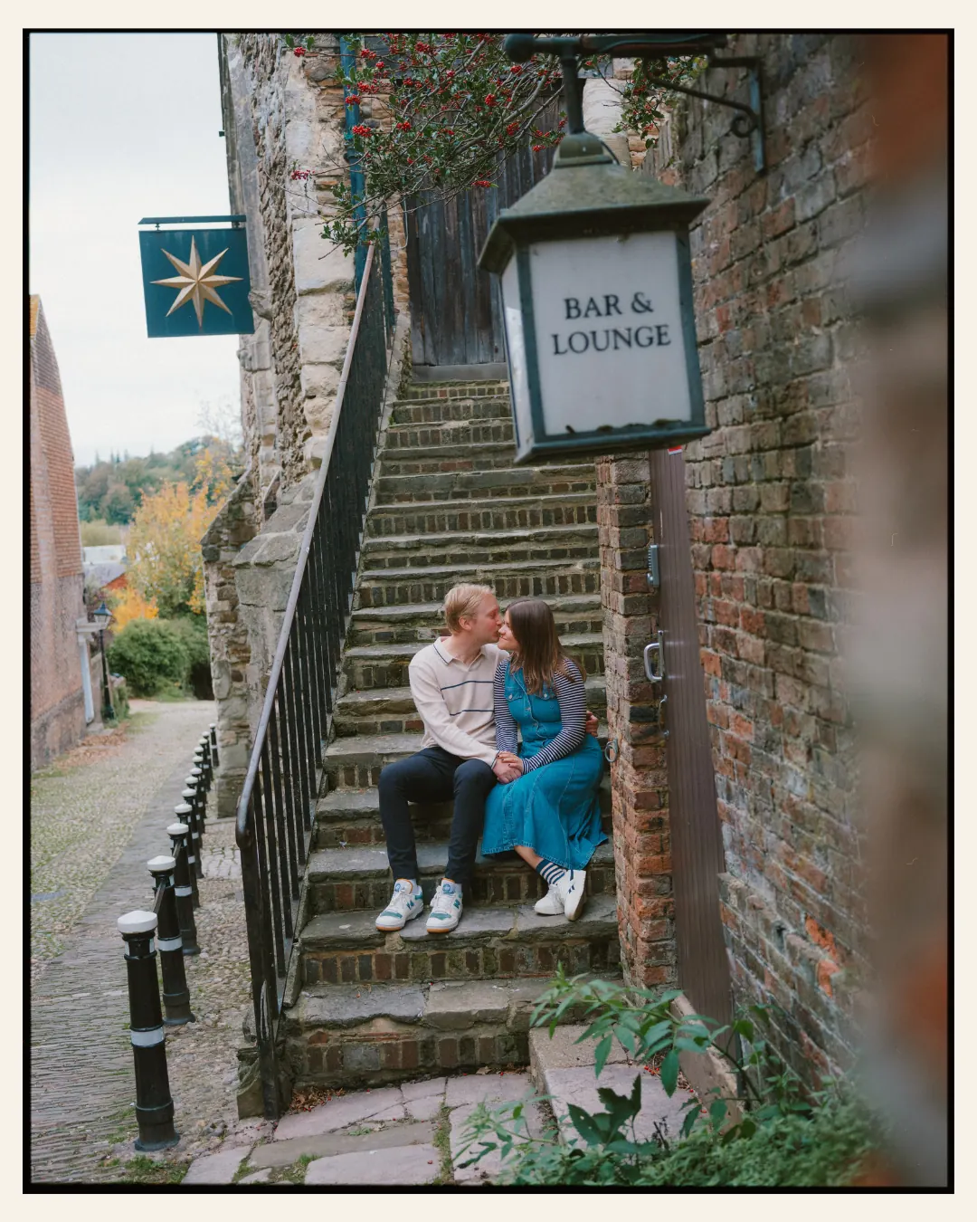 american couple sat on steps in a cobbled alley way in front of tudor building in the ancient town of rye for their engagement shoot ahead of their destination wedding