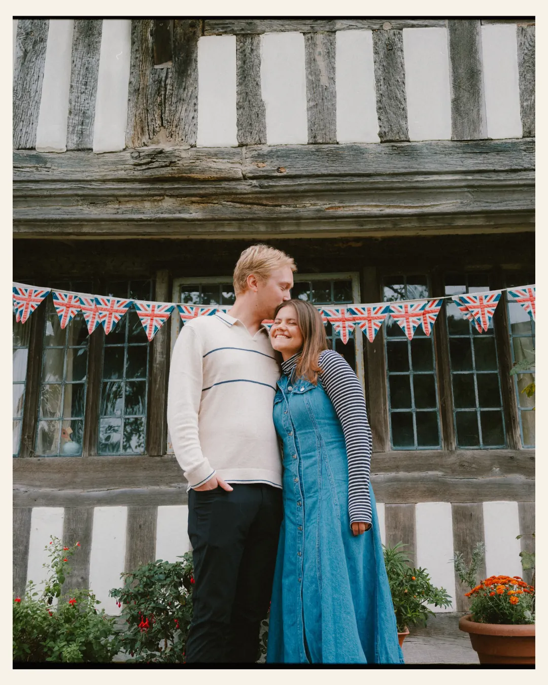 american couple standing in front of union jack bunting on an old tudor building in the ancient town of rye for their engagement shoot ahead of their destination wedding