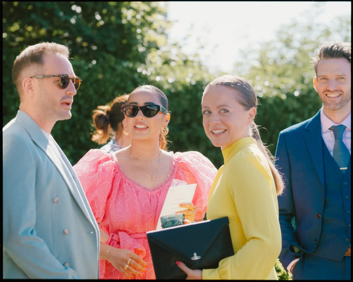 Four people dressed in colorful, stylish outfits stand outdoors, smiling and talking on a sunny day. One woman holds a clutch and another wears sunglasses and a bright pink dress. Trees and blue sky are in the background.