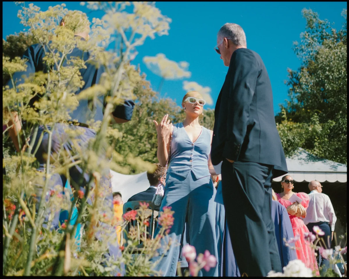 People in colorful clothing stand and talk at an outdoor event on a sunny day. A woman in sunglasses gestures while speaking to a man in a dark suit. Wildflowers and tents are visible under a clear blue sky.