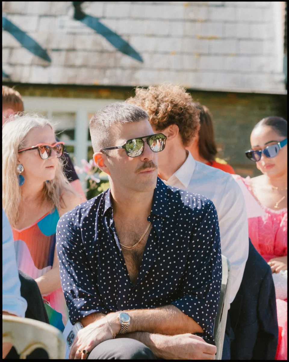 A man with short hair, a mustache, sunglasses, and a navy polka dot shirt sits outdoors among others at a sunny event. People around him wear colorful clothes and sunglasses. The background shows a stone building and sunlight.
