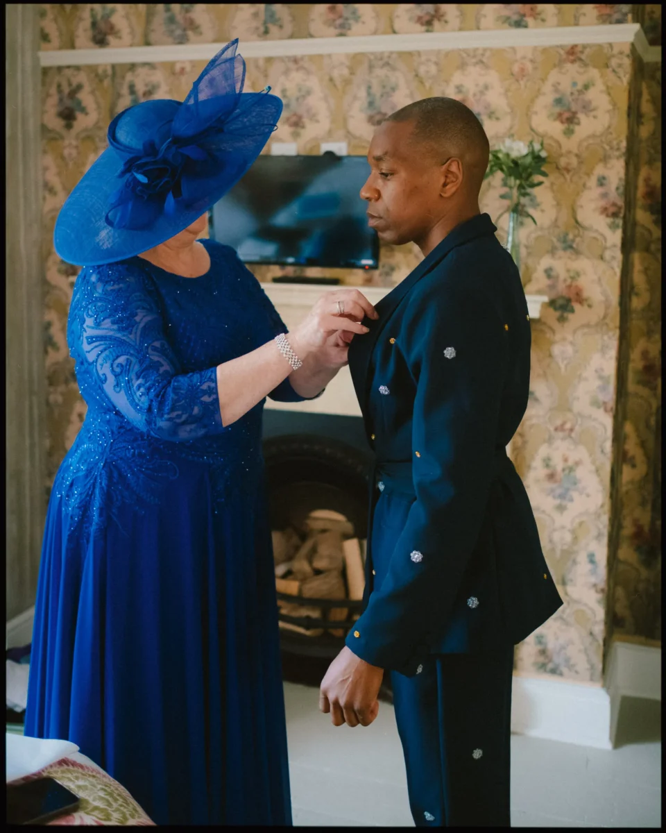 A woman in a royal blue dress and wide-brimmed hat adjusts the lapel of a mans dark suit. They stand in a warmly decorated room with floral wallpaper and a fireplace in the background.