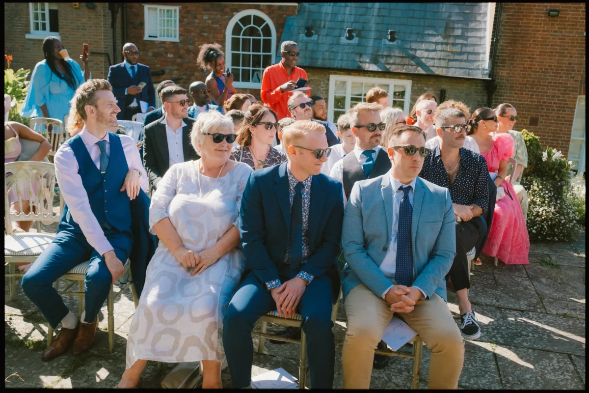 A group of people dressed in formal and semi-formal attire sit outdoors on chairs, wearing sunglasses, at a sunlit event in front of a brick building with arched windows.