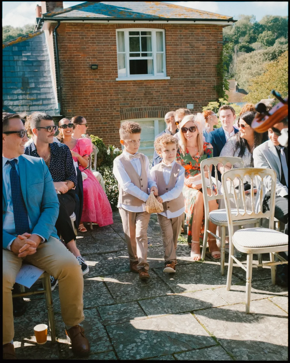 Two young boys dressed in beige suits walk down an outdoor aisle, scattering flower petals from a basket, surrounded by seated guests at a sunny wedding ceremony.