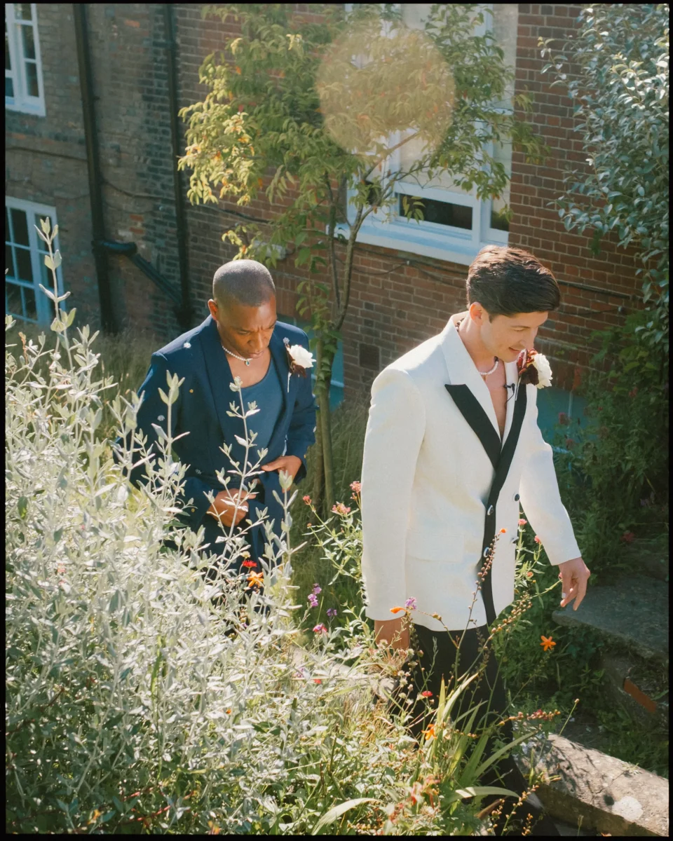 Two men dressed in suits, one in navy blue and the other in a white jacket with black accents, walk up a garden path surrounded by greenery and flowers, with sunlight filtering through trees.