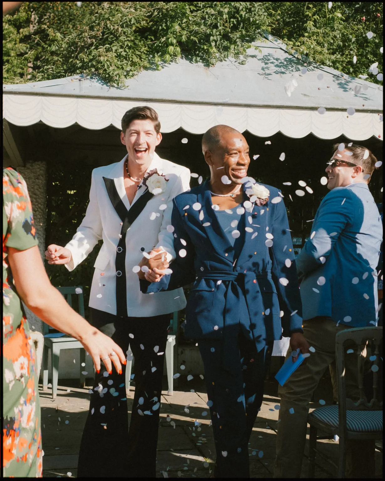 Two grooms, smiling and holding hands, walk down an aisle outdoors as guests toss white confetti. One groom wears a white tuxedo jacket, the other a navy suit. It’s a sunny day, and people rejoice around them.