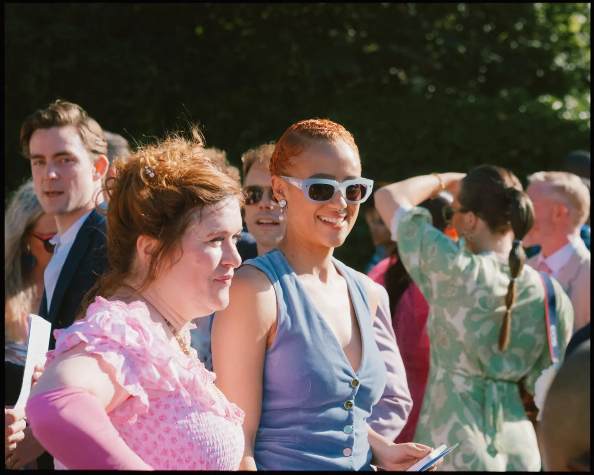 A group of people outdoors dressed in colorful, formal attire. A woman in pink and another in a blue dress with white sunglasses are smiling in the foreground. Others converse and mingle in the background.