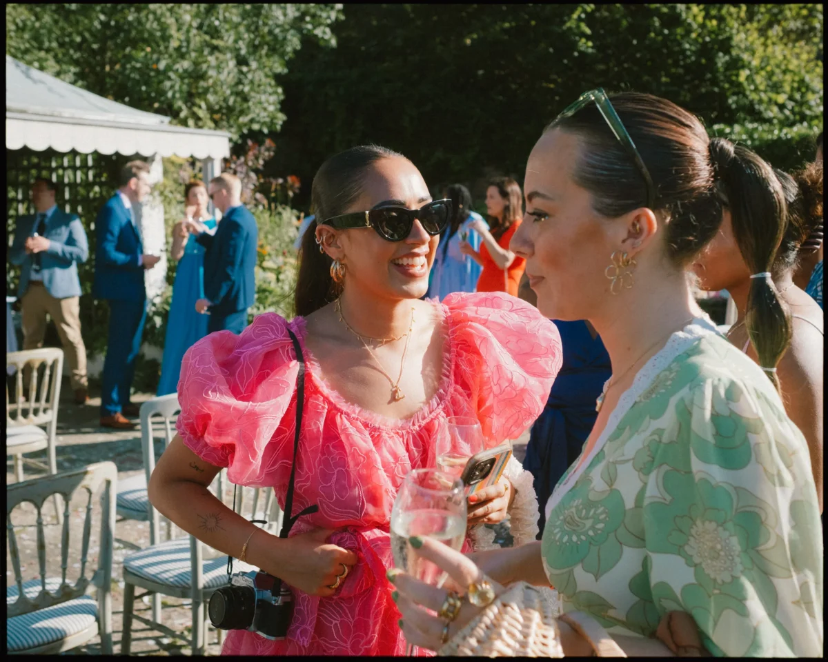 Two women smiling and talking at an outdoor event, both dressed in colorful, stylish outfits. One wears sunglasses and a pink dress, holding a camera; the other wears a floral green-and-white dress, holding a glass of water.