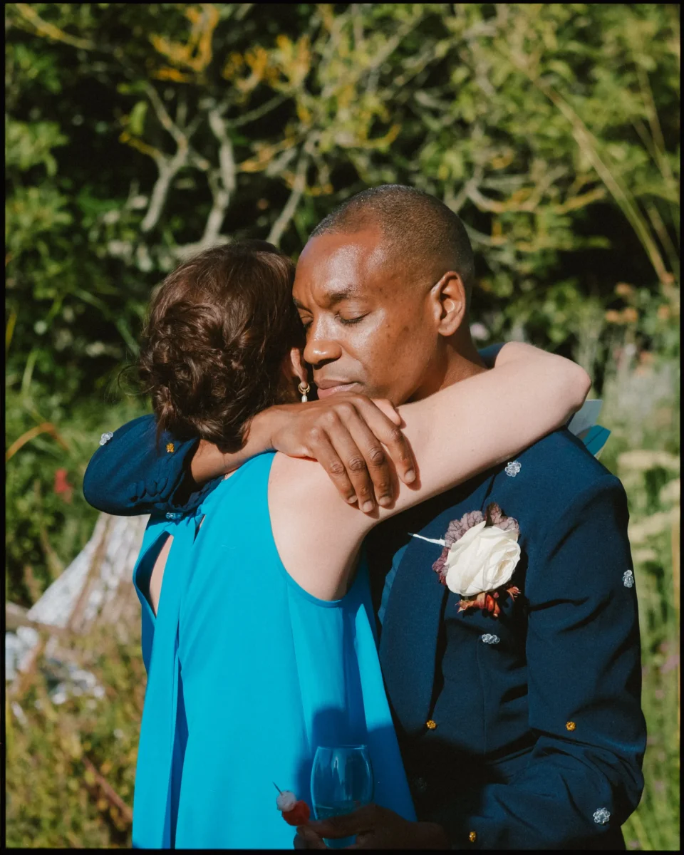 A man in a navy suit with a white rose boutonniere embraces a woman in a bright blue dress outdoors. They have their eyes closed, sharing a warm, emotional hug in a sunlit garden setting.