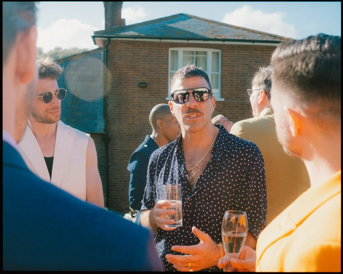 A man in a black polka-dot shirt and sunglasses holds a drink while talking with others at an outdoor gathering on a sunny day, with a brick house in the background.