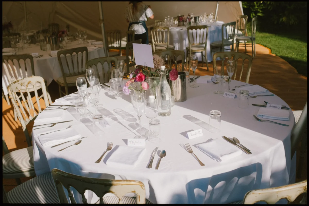 A round table set for a formal event with white tablecloth, glassware, cutlery, napkins, and a floral centerpiece. Place cards are arranged at each seat. More similarly set tables and a person are visible in the background.