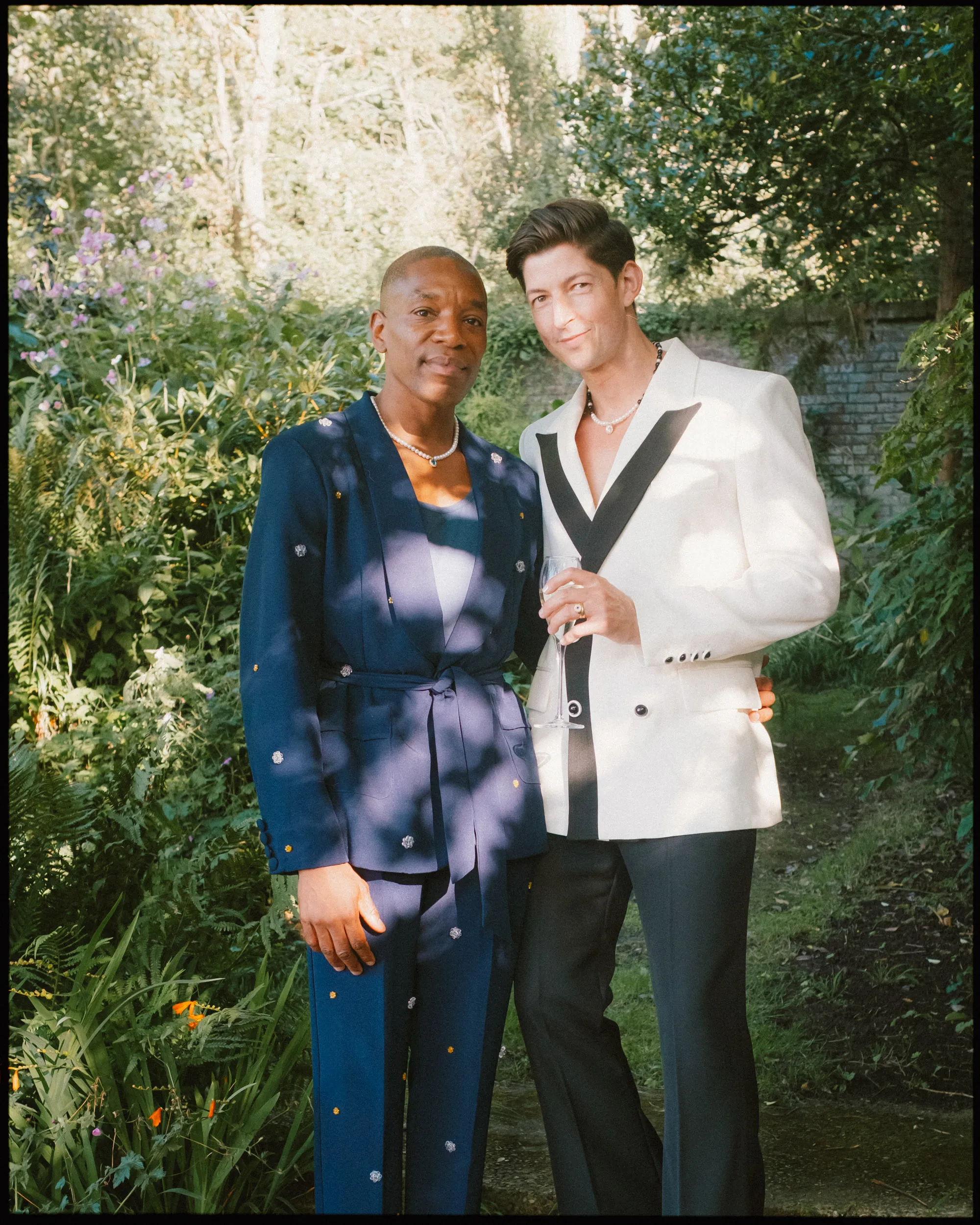 Two people stand close together in a lush garden. One wears a dark blue suit with small white dots, and the other wears a white suit with black trim, holding a glass. Sunlight filters through the trees in the background.