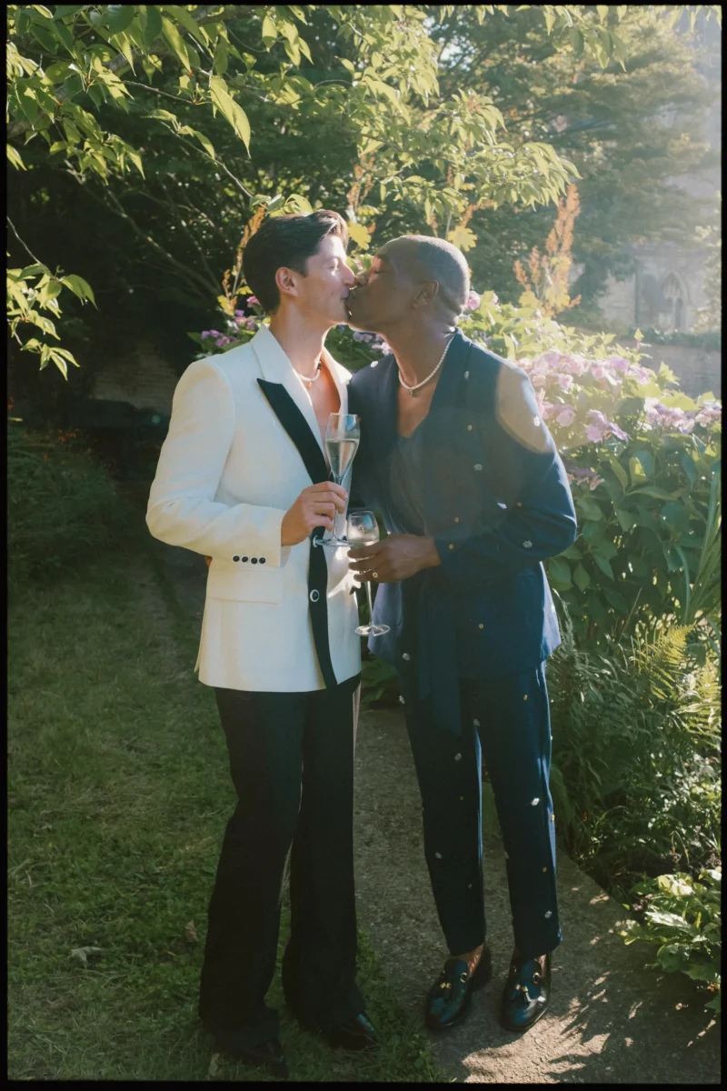 Two people dressed formally, one in a white blazer and the other in a navy suit, share a kiss while holding champagne glasses in a sunlit garden with greenery and purple flowers in the background.