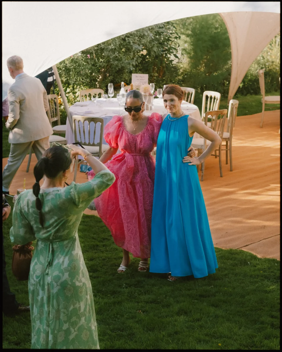 Two women in bright dresses pose for a photo at an outdoor event; one wears a pink gown and sunglasses, the other a blue dress. Another woman in green takes their picture. Empty tables and chairs are visible in the background.