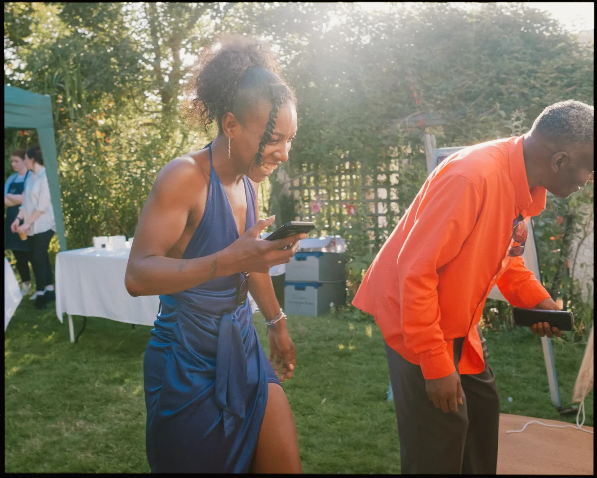 A woman in a blue dress and a man in an orange shirt smile and laugh while holding phones outdoors, with greenery and people in the background at what appears to be a garden party.