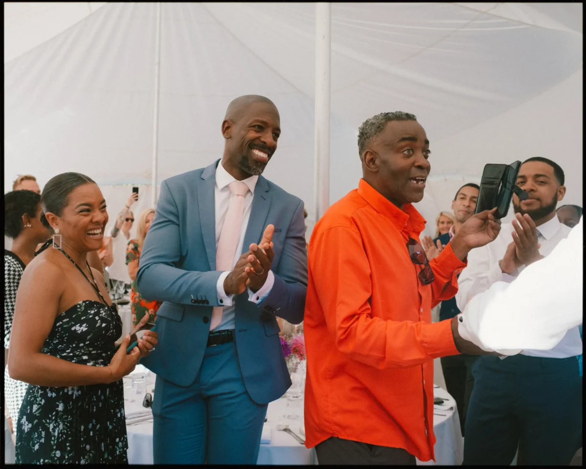 A group of people dressed in semiformal attire smile and clap at an indoor event. A man in a blue suit and a woman in a black dress stand beside a man in a bright red shirt, all appearing happy and engaged.