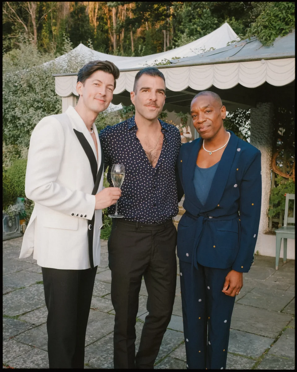 Three people stand outdoors dressed in formal attire. One holds a champagne glass, and all are posing for the photo, with greenery and a white canopy tent in the background.