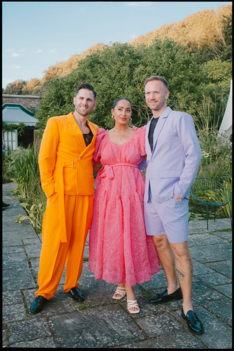 Three people pose outdoors in bright, colorful outfits: one in an orange suit, one in a pink dress, and one in a lilac suit with shorts. They stand on a stone patio with greenery and trees in the background.