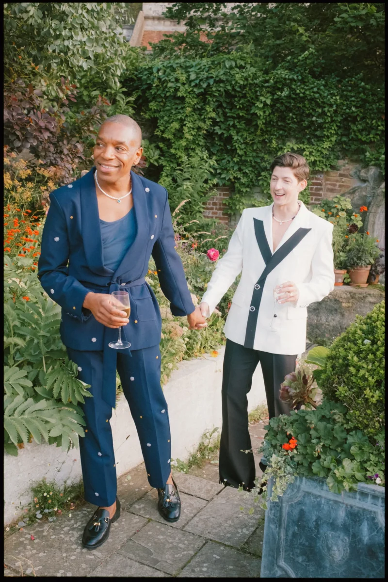 Two people dressed in stylish suits, one in navy blue and the other in white and black, hold hands and smile while standing in a lush garden, each holding a drink.