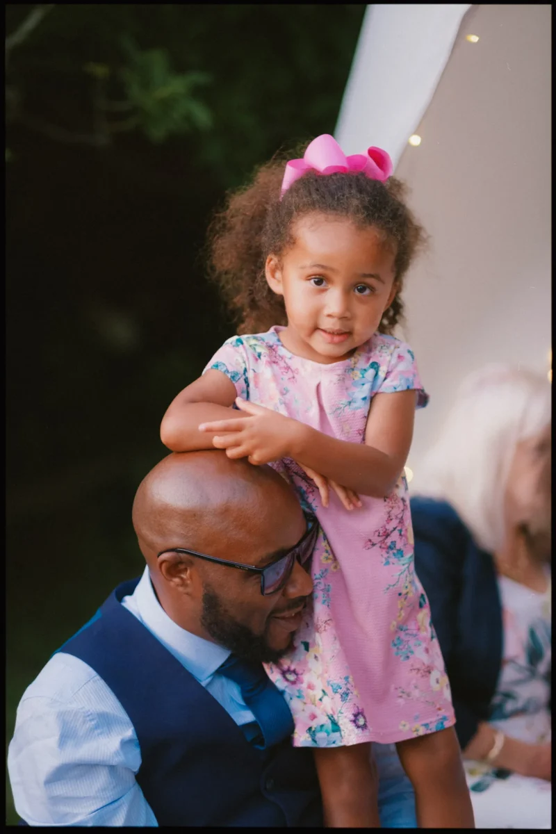 A young girl with curly hair and a pink bow stands smiling in a floral dress, resting her arms on the shoulder of a bearded man in glasses and a suit vest, with blurred people and greenery in the background.