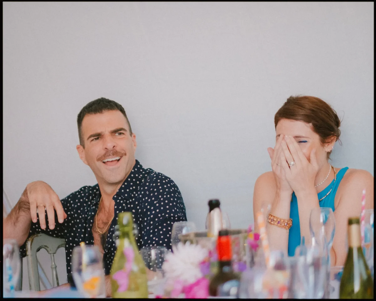 Two people sit at a table decorated with flowers and bottles. The man on the left smiles and looks amused, while the woman on the right, wearing blue, laughs with her hand covering her face.