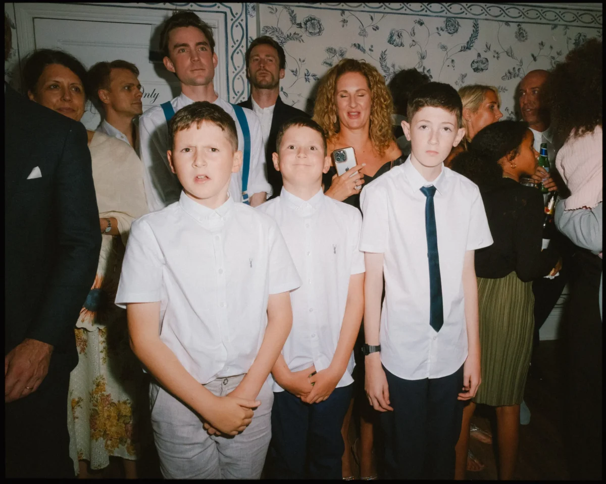 A group of people dressed formally at a gathering; three boys in white shirts stand at the front, with adults and children in the background against a patterned wallpaper.