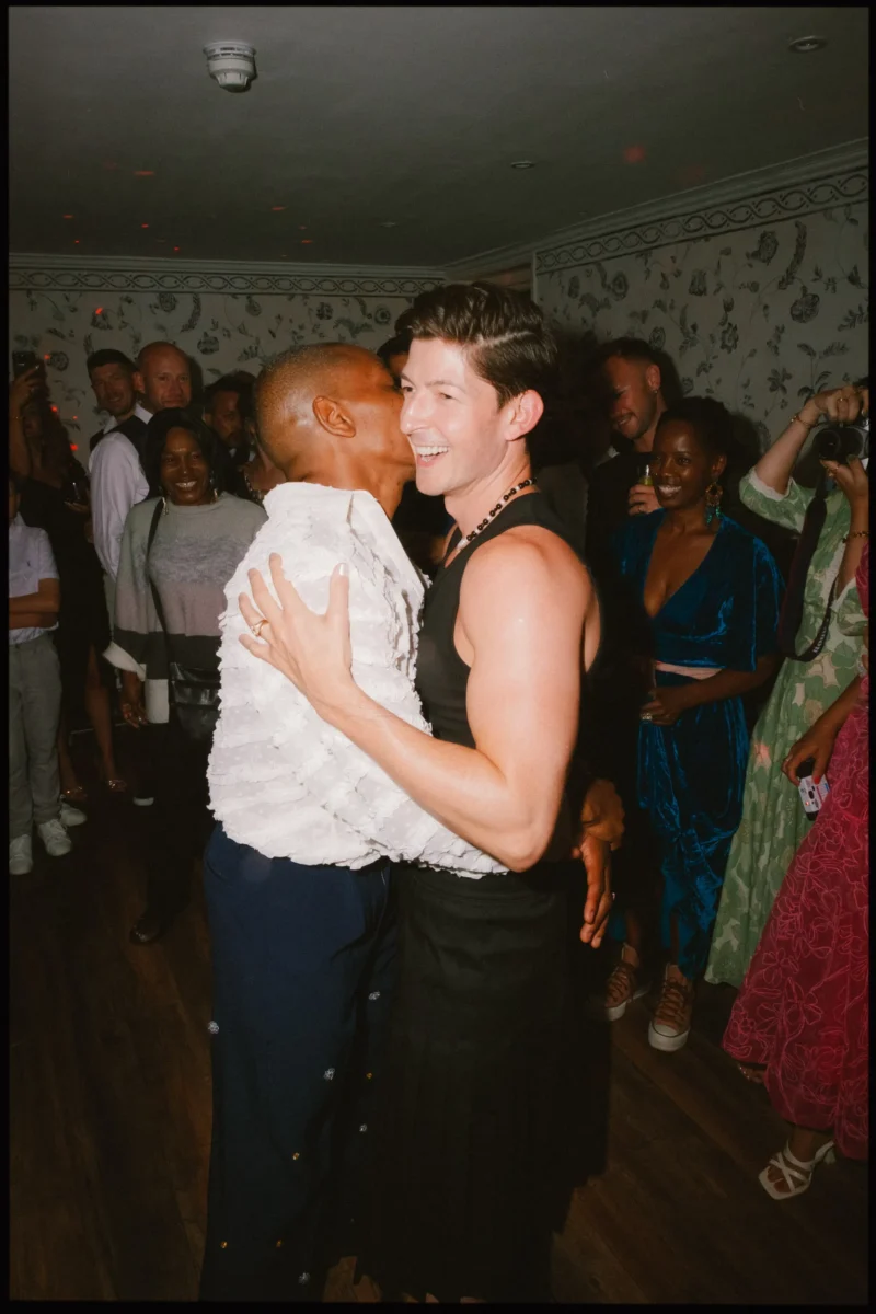 Two people joyfully embrace and smile in the center of a crowded room. The group around them is watching, some smiling and taking photos. The setting appears celebratory, with casual and colorful outfits.