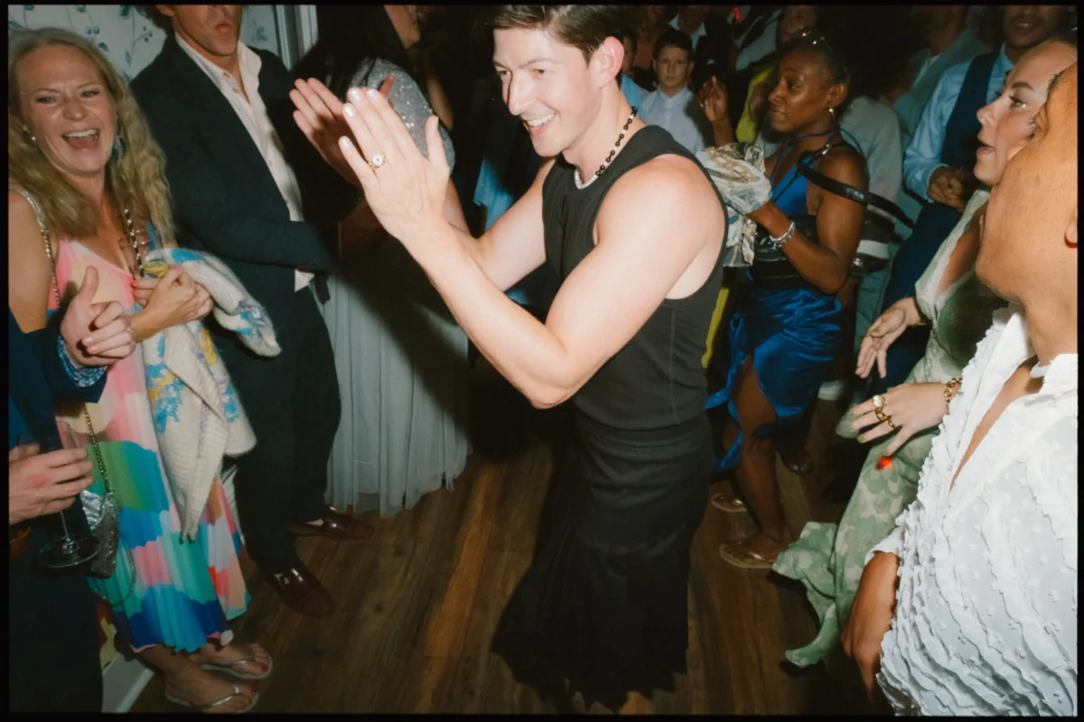 A person in a black sleeveless top and skirt claps joyfully while dancing in a lively crowd. People around them smile, laugh, and cheer, dressed in colorful and formal attire on a wooden floor.