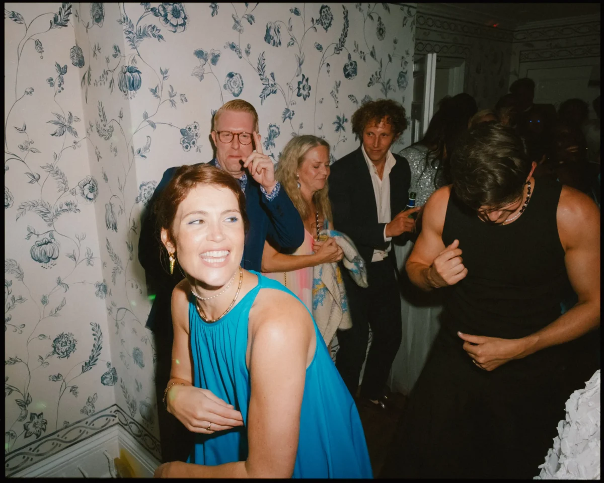 A group of people dance and smile at a party in a room with floral wallpaper. A woman in a blue dress is in the foreground, while others mingle and enjoy themselves in the background.