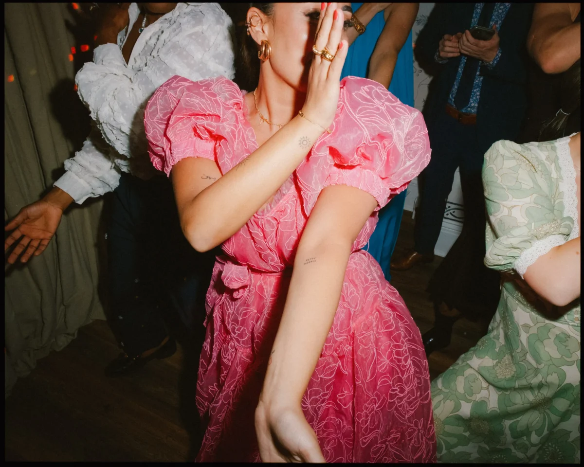 A woman in a bright pink, puff-sleeved dress dances energetically, partially covering her face with her arm. Other people in formal attire dance around her in a lively, indoor setting.