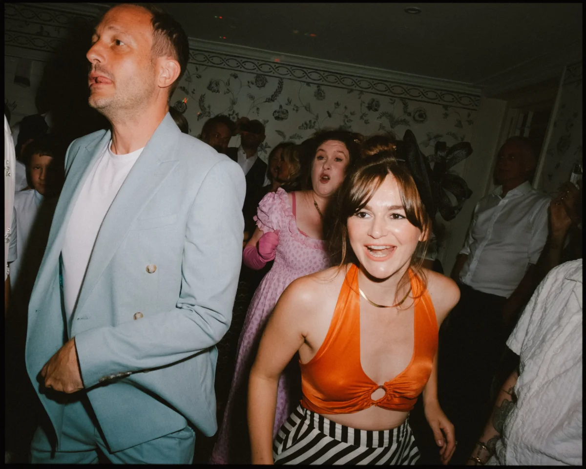 People dancing at a lively indoor party. A woman in an orange top and striped pants smiles in the foreground, with a man in a light blue suit and a woman in a pink dress behind her. The room is crowded and festively decorated.