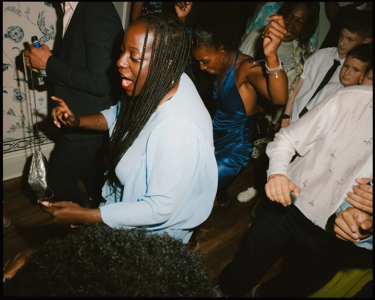 A group of people dance closely together at a lively indoor party. A woman in a light blue outfit is in the foreground, surrounded by others in colorful dresses and shirts, all enjoying the music.