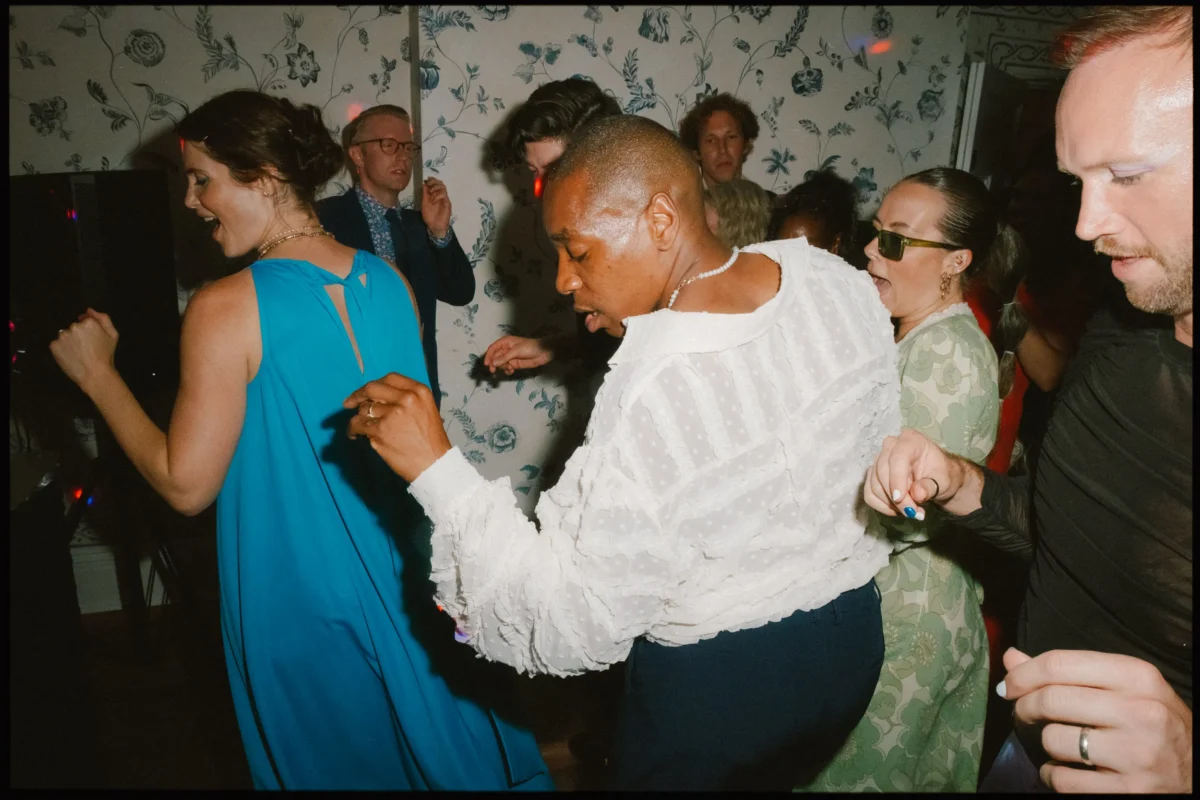 A group of people enthusiastically dancing indoors at a party, dressed in colorful and formal attire, with patterned wallpaper and dim lighting in the background.